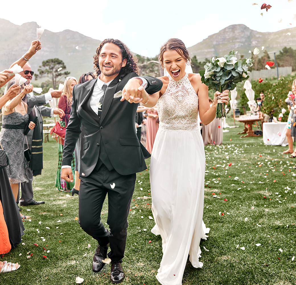 Man wearing suit and woman wearing wedding dress walking up grassy wedding aisle while having petals thrown at them by guests